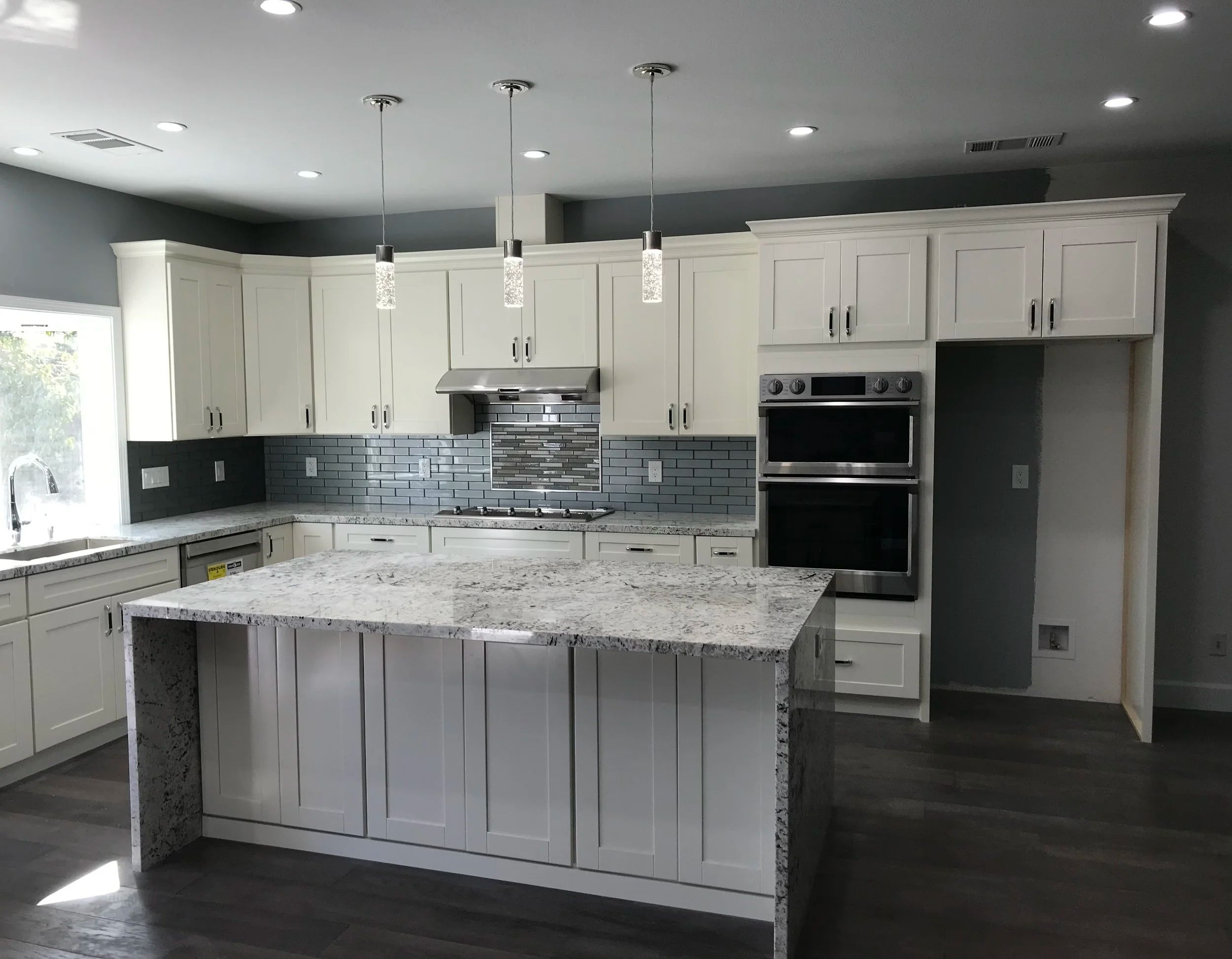 Remodeled kitchen with white shaker cabinets, a granite island, three glass pendant lights, a grey tile backsplash and stainless double wall ovens.