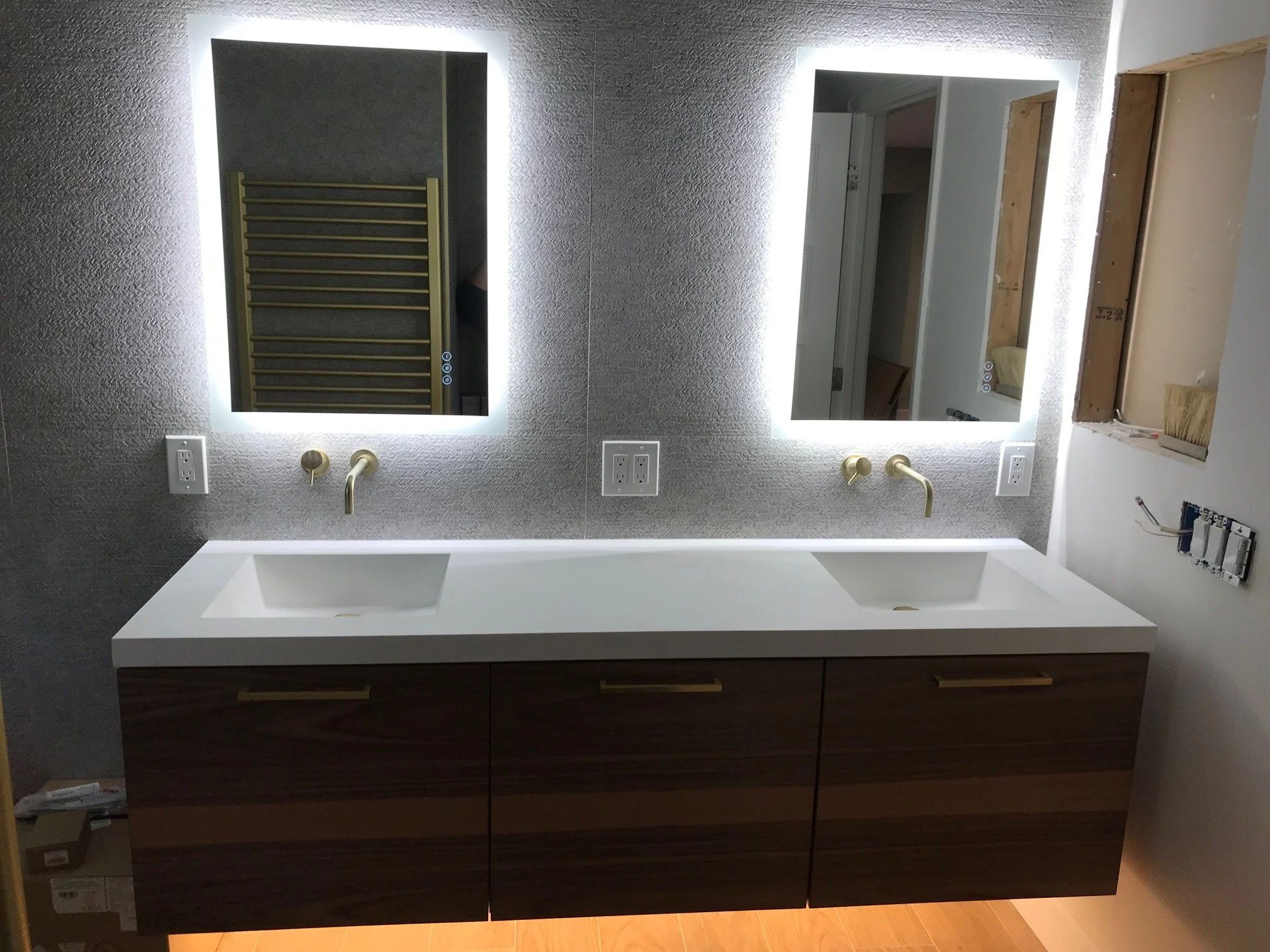 Bathroom mid-remodel with a floating walnut double vanity, backlit mirrors and wall-mounted brass faucets, with wall finishes still in progress.