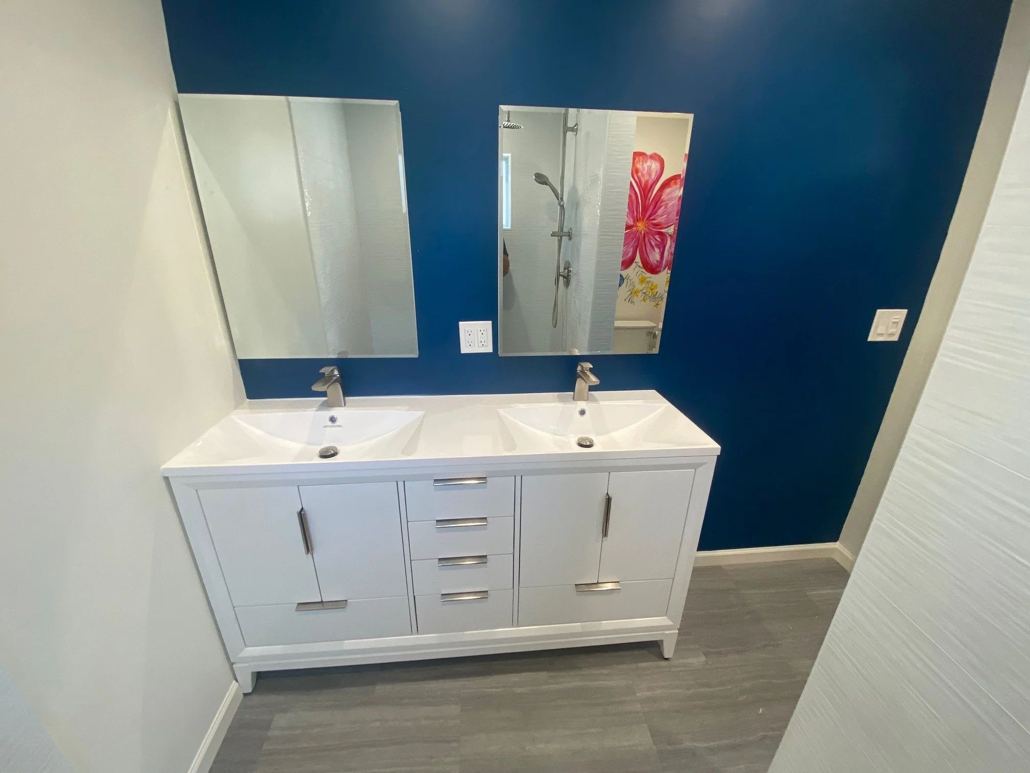 Remodeled bathroom with a white double vanity, two mirrors, a deep navy accent wall and wood-look plank flooring.