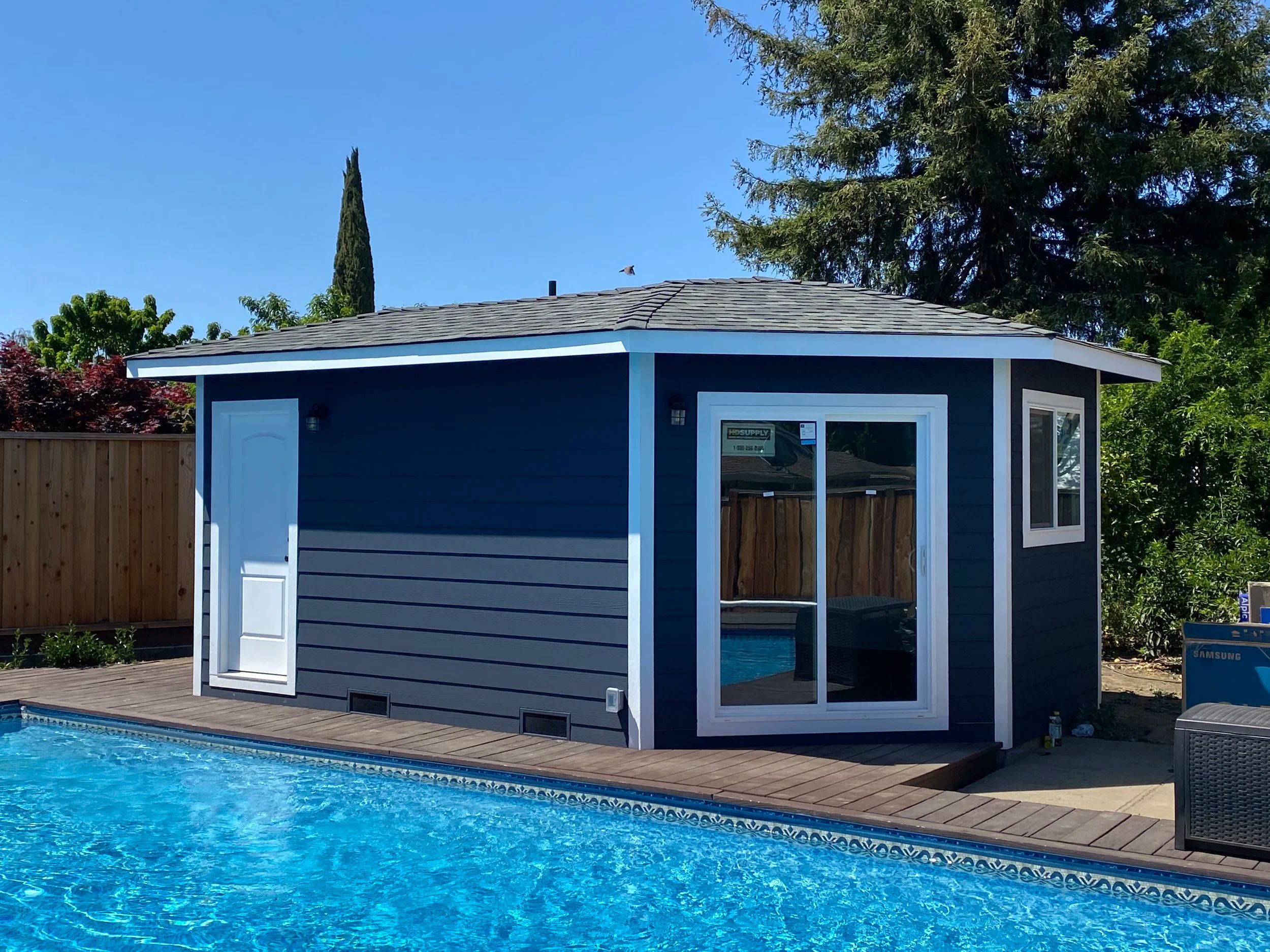 Detached poolside building with navy lap siding, a hipped roof and a glazed door, built beside a swimming pool.