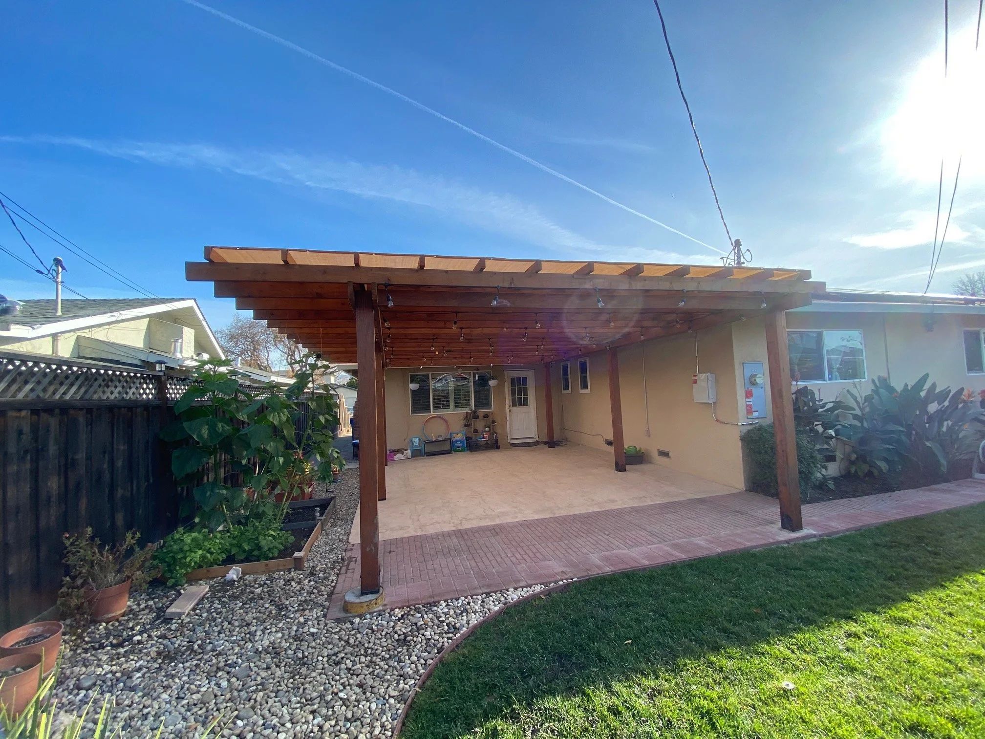 Redwood patio cover built across the back of a house, over a brick and concrete patio, strung with festoon lighting.